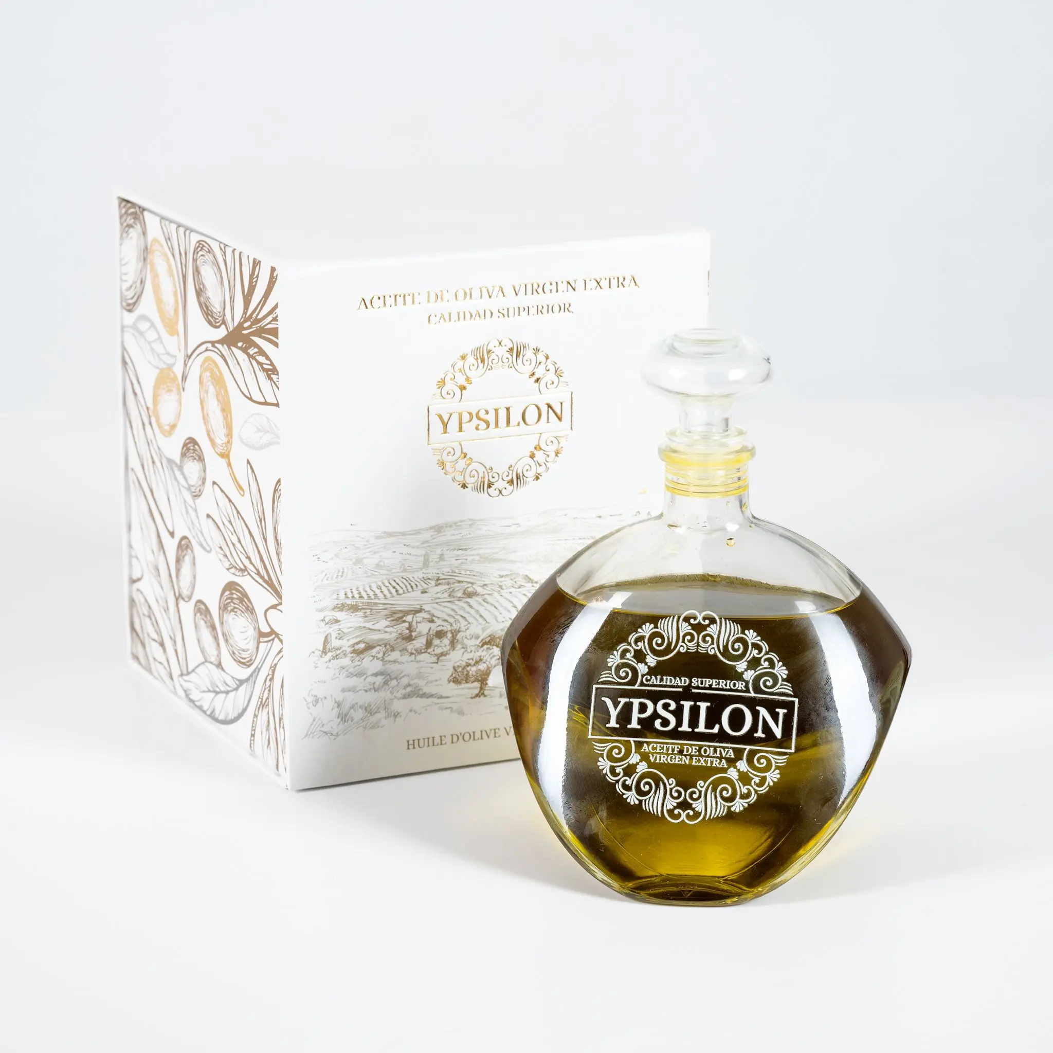 Hera Premium Bio-Olivenöl Picual (Ypsilon Calidad Superior) in einer runden Karaffe neben der geöffneten weißen Geschenkschachtel mit goldenen Verzierungen und Olivenzweig-Motiven, auf weißem Studiohintergrund.