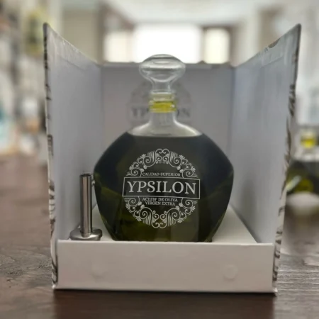 Hera Premium Bio-Olivenöl (Picual) in der Ypsilon-Karaffe, präsentiert in einem weißen Schachteleinsatz neben einem mitgelieferten Edelstahl-Gießausgießer, mit unscharfem Delikatessengeschäft-Hintergrund.