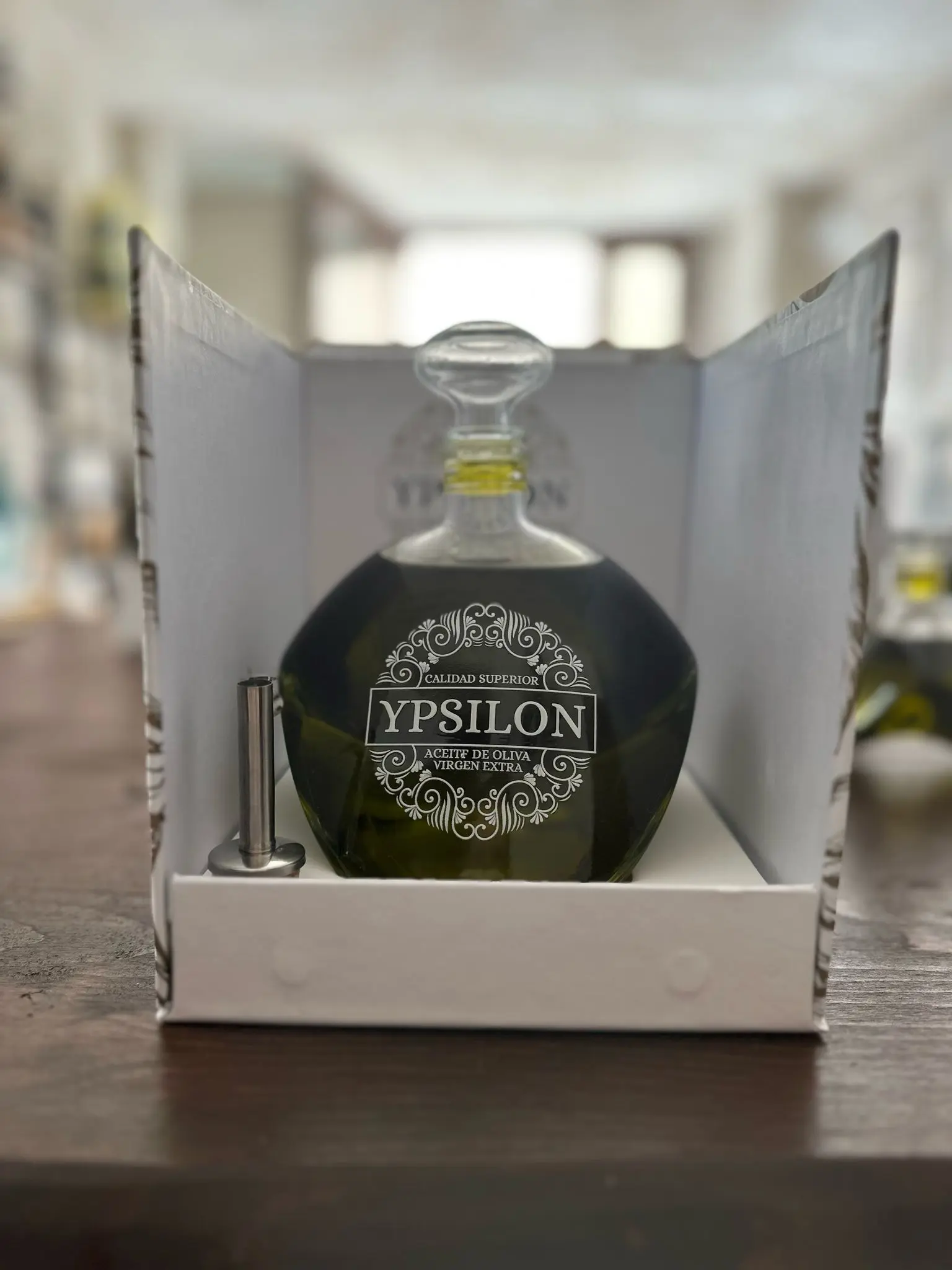 hera-olivenoel-picual-ypsilon-karaffe-im-geschenkkarton Hera Premium Bio-Olivenöl (Picual) in der Ypsilon-Karaffe, präsentiert in einem weißen Schachteleinsatz neben einem mitgelieferten Edelstahl-Gießausgießer, mit unscharfem Delikatessengeschäft-Hintergrund.