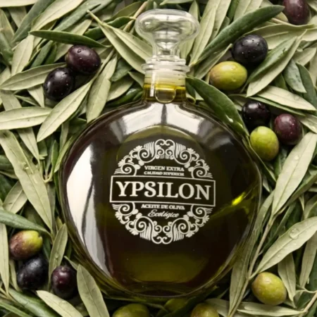 Top-Down-Foto der markanten Hera Premium Bio-Olivenöl-Karaffe (Ypsilon), die auf einem üppigen Bett aus frischen grünen Olivenblättern und grünen sowie reifen violetten Olivenfrüchten liegt.