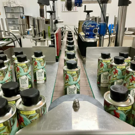 Zahlreiche Tropicual de Sierra Olivenöldosen auf einem metallischen Förderband in einer modernen Abfüllanlage. Die Dosen tragen zusätzliche 