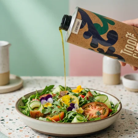 Aus der Tropicual Everyday Olivenöl-Dose wird frisches, goldgrünes Öl über einen bunten Salat mit Tomaten, Gurken und essbaren Blüten auf einem Terrazzo-Tisch gegossen.