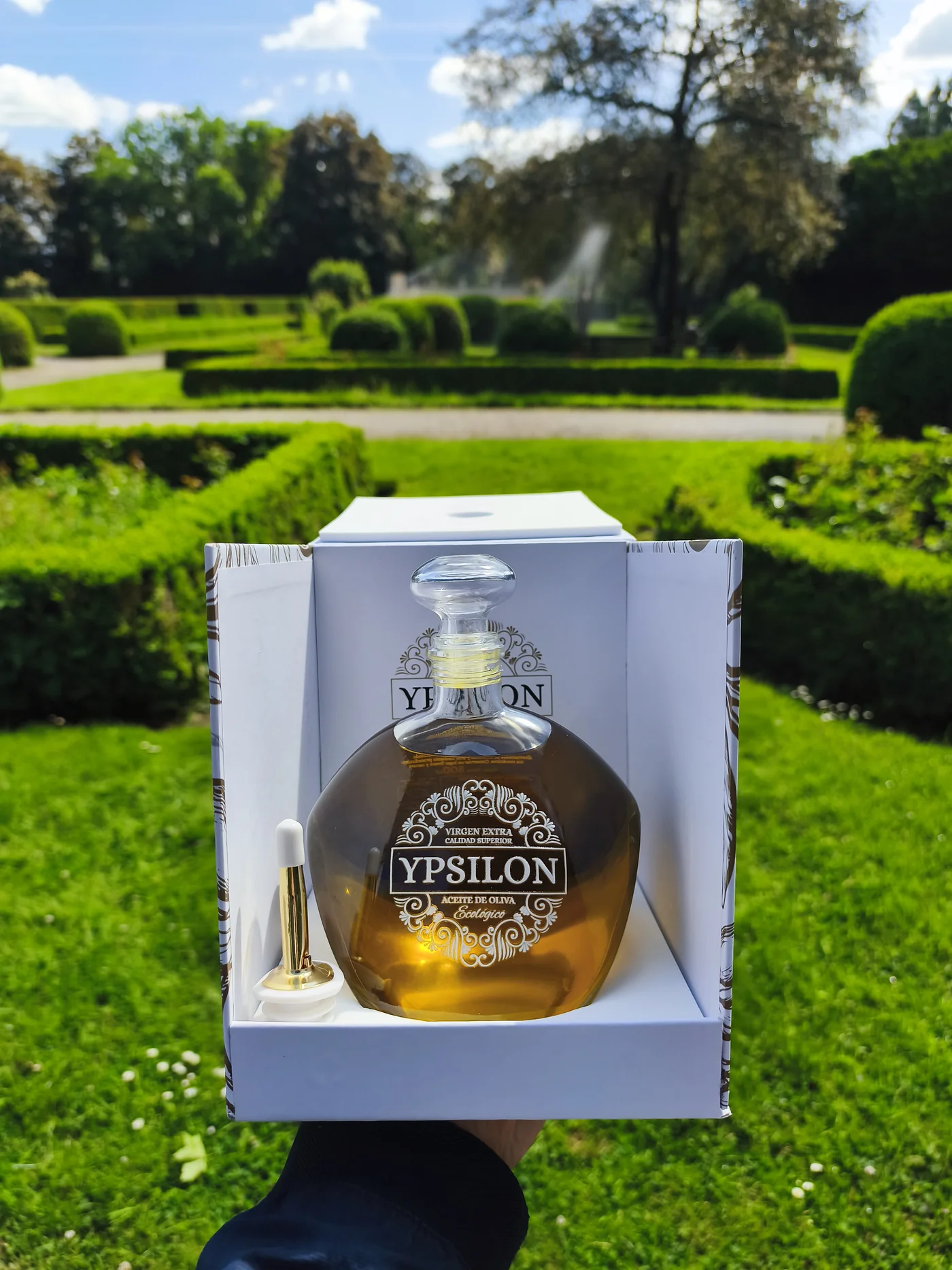 Hand hält geöffnete Geschenkbox mit einer Flasche Hera Ypsilon Olivenöl vor einem gepflegten Schlossgarten mit Hecken und Springbrunnen im Hintergrund.